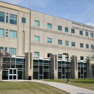 A four-storey tan brick building with large windows on the bottom floor and a concrete path leading through a grassy lawn to the front door.